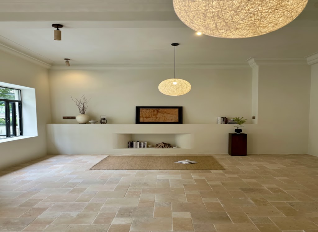 Empty living room with beige travertine tile flooring, minimalist built-in shelving, and natural light from window.png
