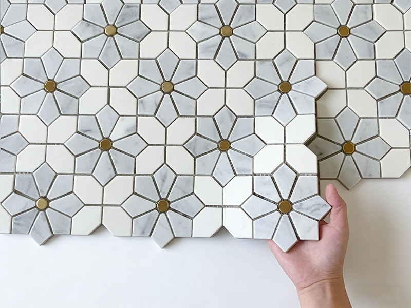 Grey and white flower-patterned stone mosaic tile with brass accents, held in hand for product display, ideal for backsplash decor.png